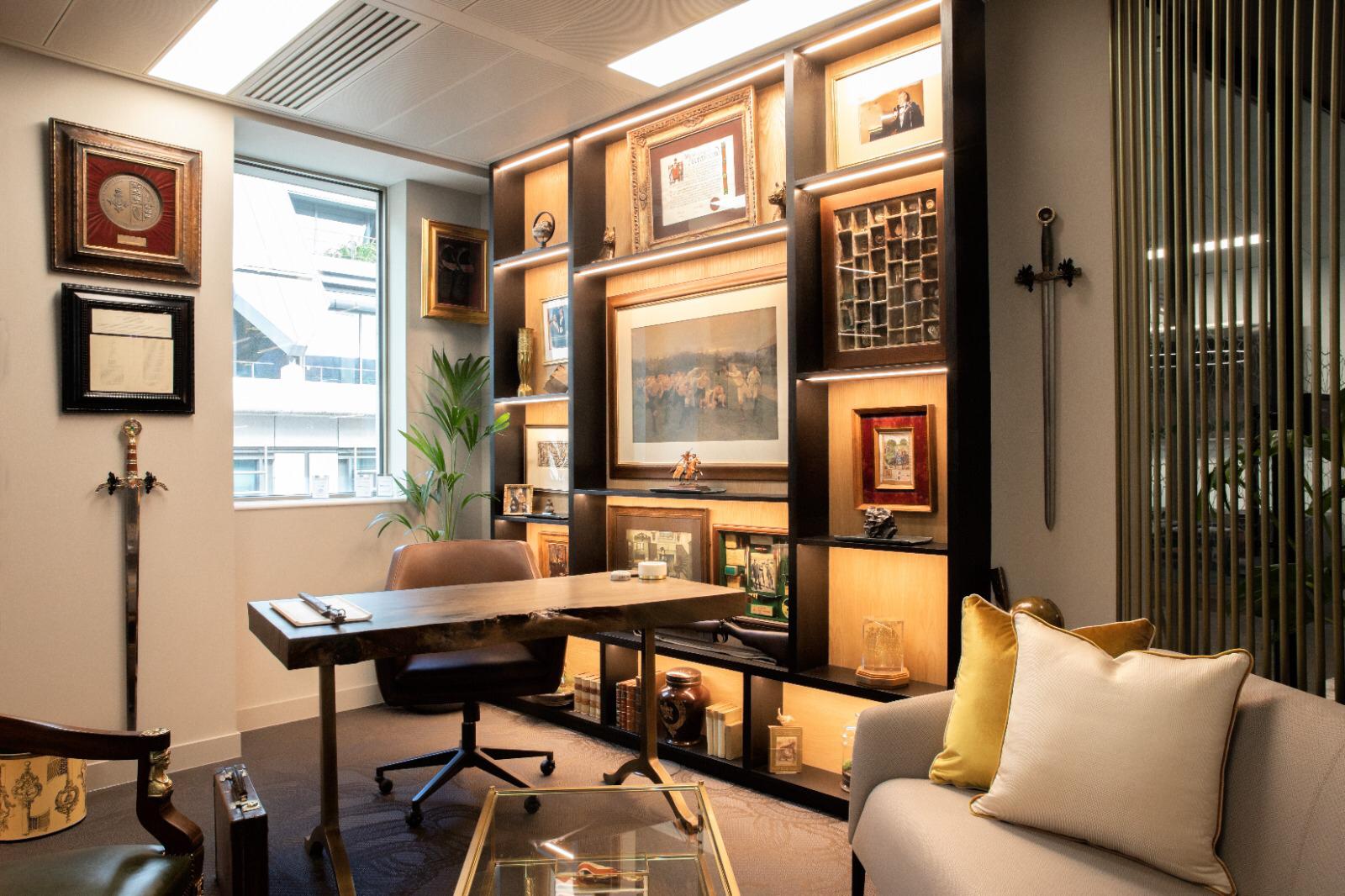 Photo of a bespoke home office with a floor-to-ceiling illuminated display shelving unit filled with framed prints and curios, a live-edge wood desk, a brown leather chair and a grey sofa.