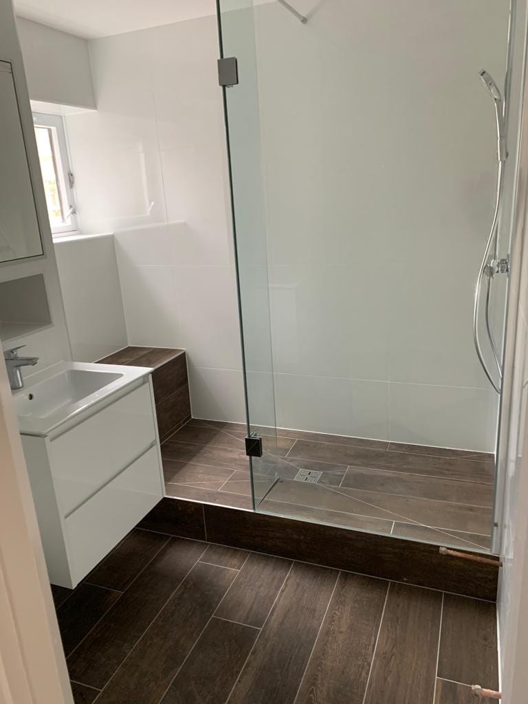 Photo of a completed bathroom with a walk-in frameless glass shower, dark wood-effect floor tiles and a white wall-hung vanity unit.