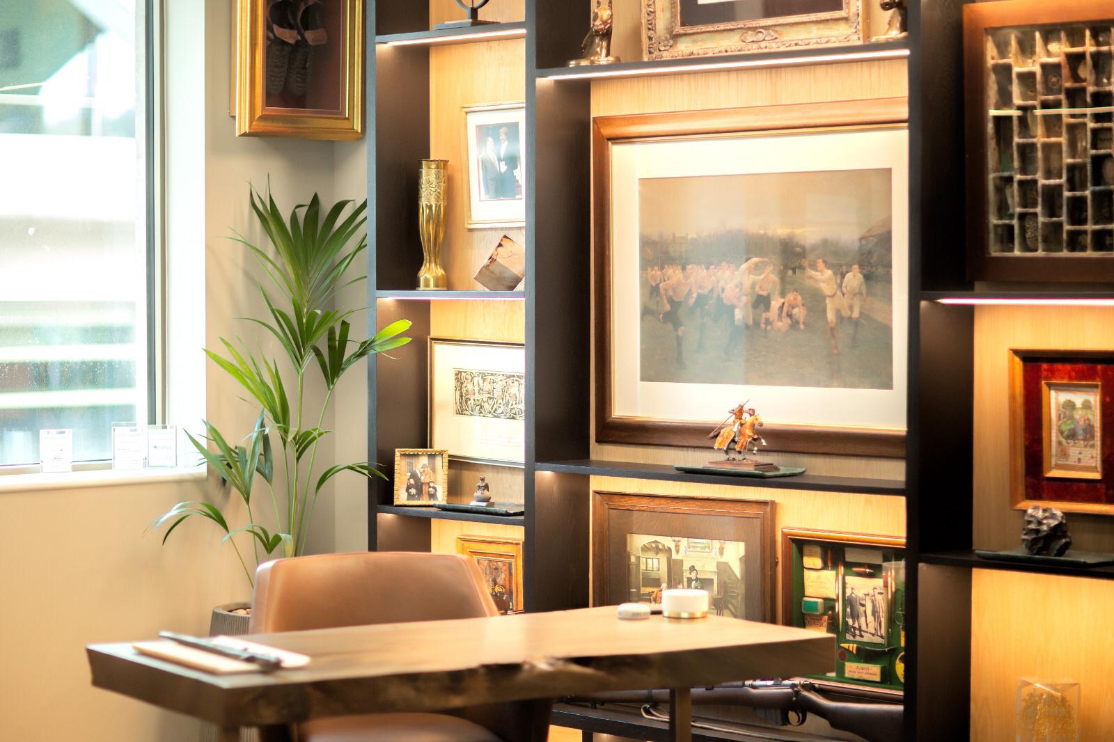 Photo of a bespoke fitted home office with a walnut-look shelving and cabinet unit displaying framed prints, apothecary jars, silver figurines and sporting memorabilia, a live-edge desk, a leather chair and a grey sofa.