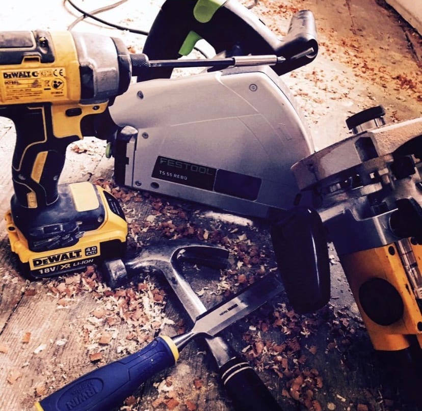 Photo of power tools — a cordless drill, a circular saw and a chisel — laid on a dusty wooden floor covered in sawdust at a construction site.