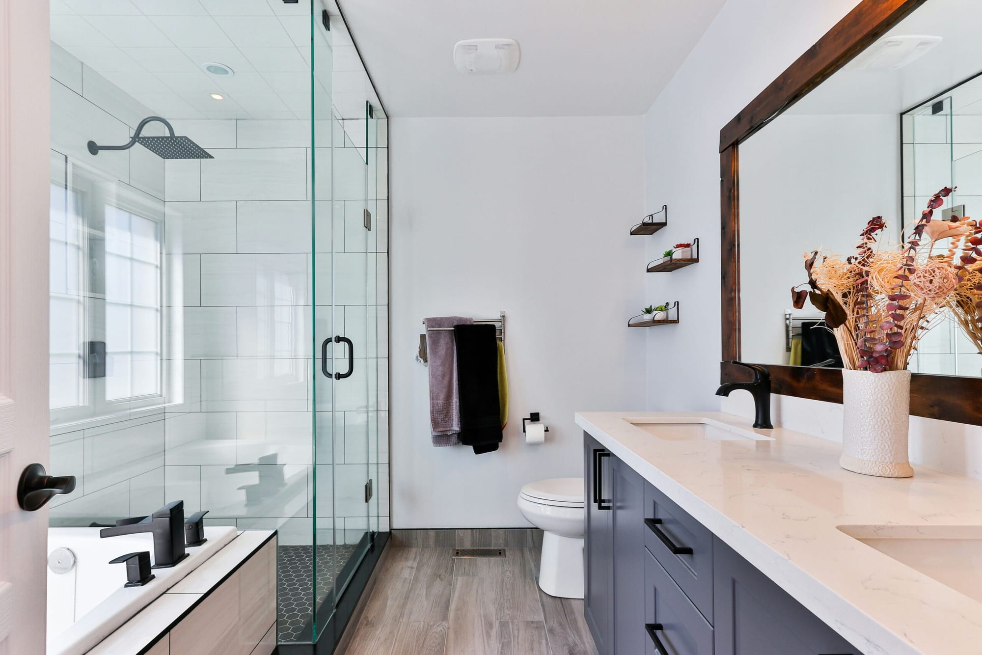 Photo of a luxury bathroom with a glass-walled walk-in shower, a freestanding white bathtub with black fixtures, a dark wood-framed mirror and a vase of dried flowers on a marble-effect vanity.