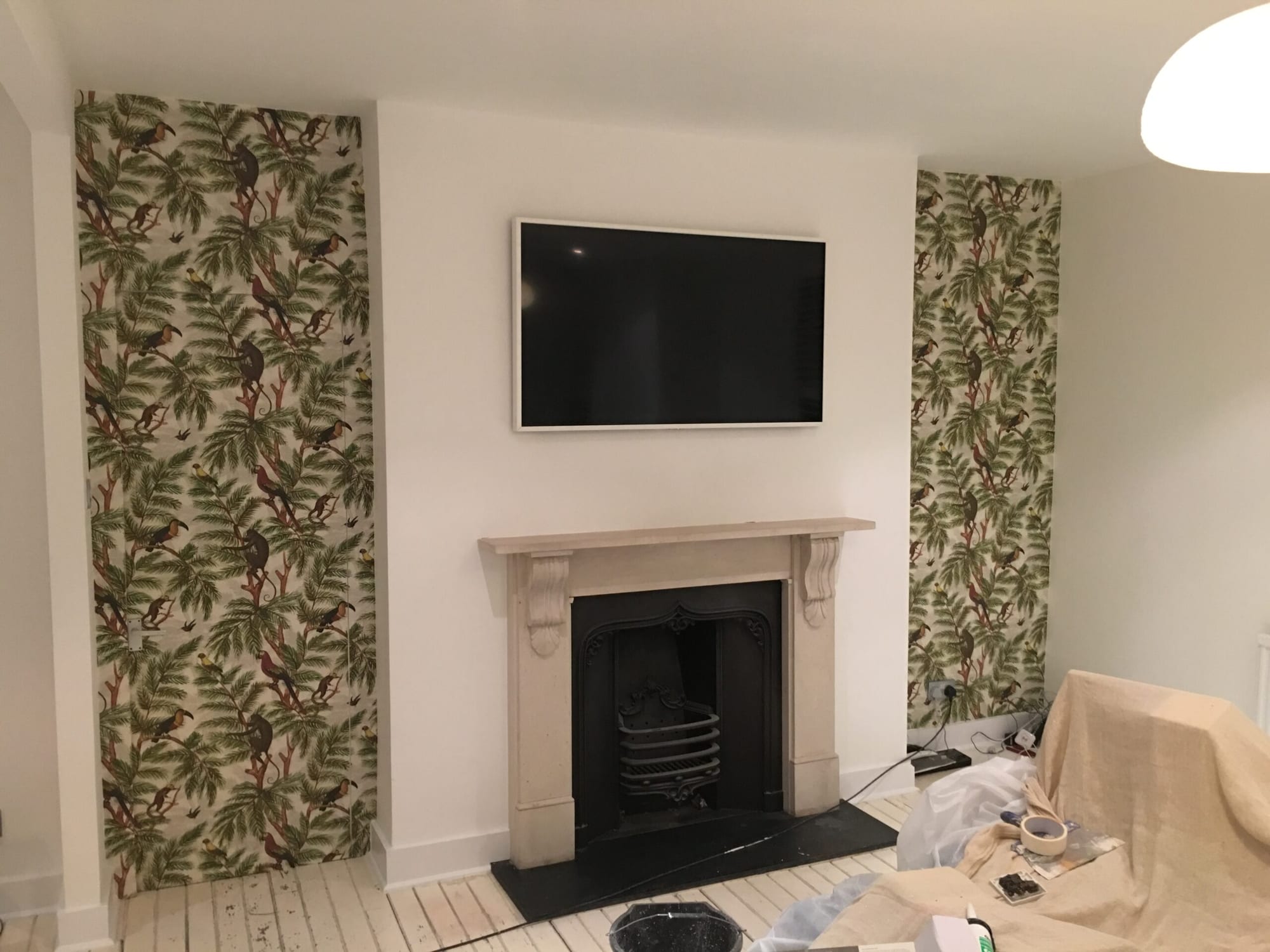 Photo of a living room mid-decoration, with jungle-print wallpaper (toucans and monkeys among palm fronds) hung either side of a stone fireplace and wall-mounted TV, dust sheets and paint tins on the floor.