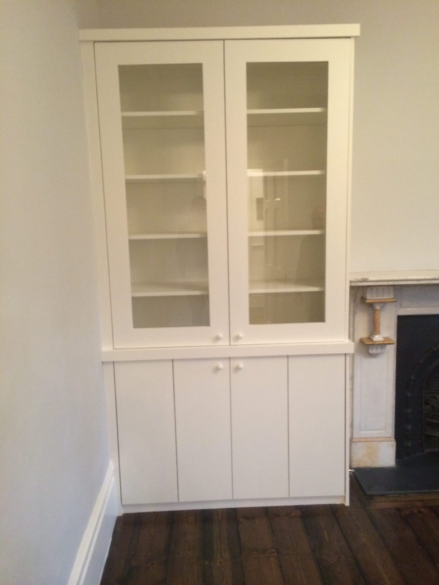 Photo of a white built-in cabinet with glazed upper doors and solid panelled lower doors, fitted into an alcove beside a period fireplace.