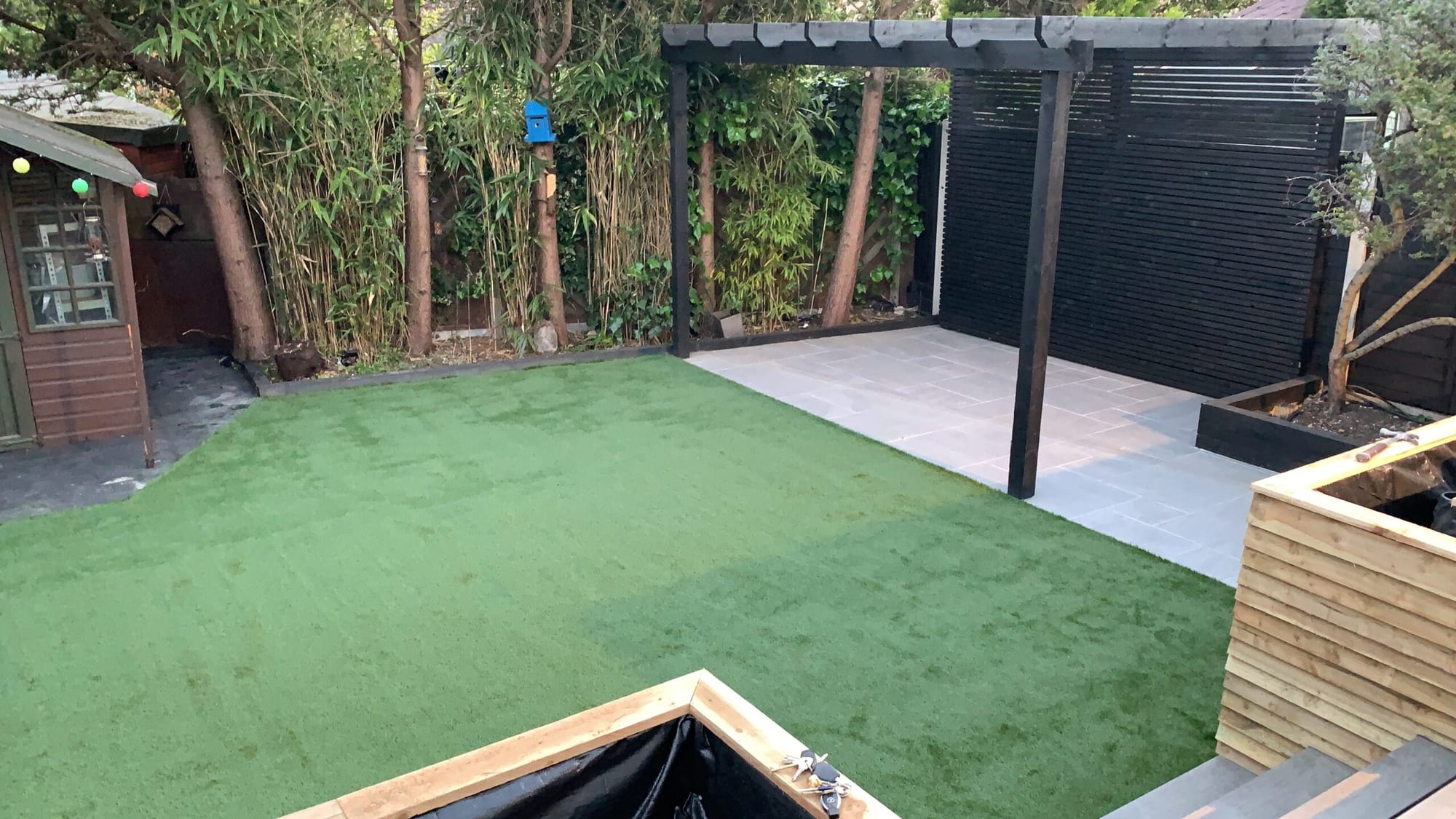 Photo of a landscaped garden with new artificial lawn, a paved patio area, a black slatted timber pergola, bamboo planting and a garden shed.
