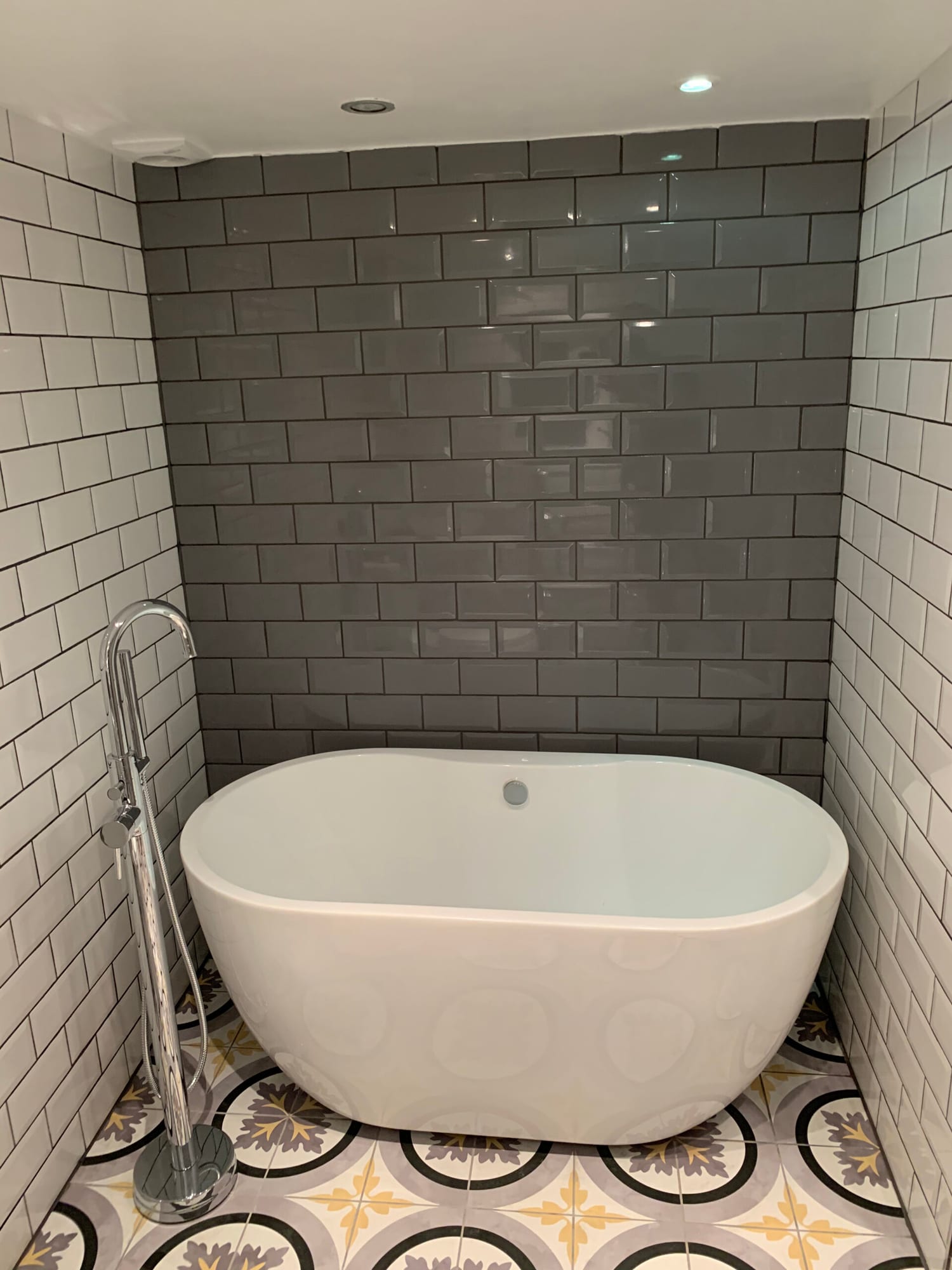 Photo of a freestanding oval bathtub with a floor-standing tap, set against a charcoal-grey metro tile splashback and patterned floor tiles.