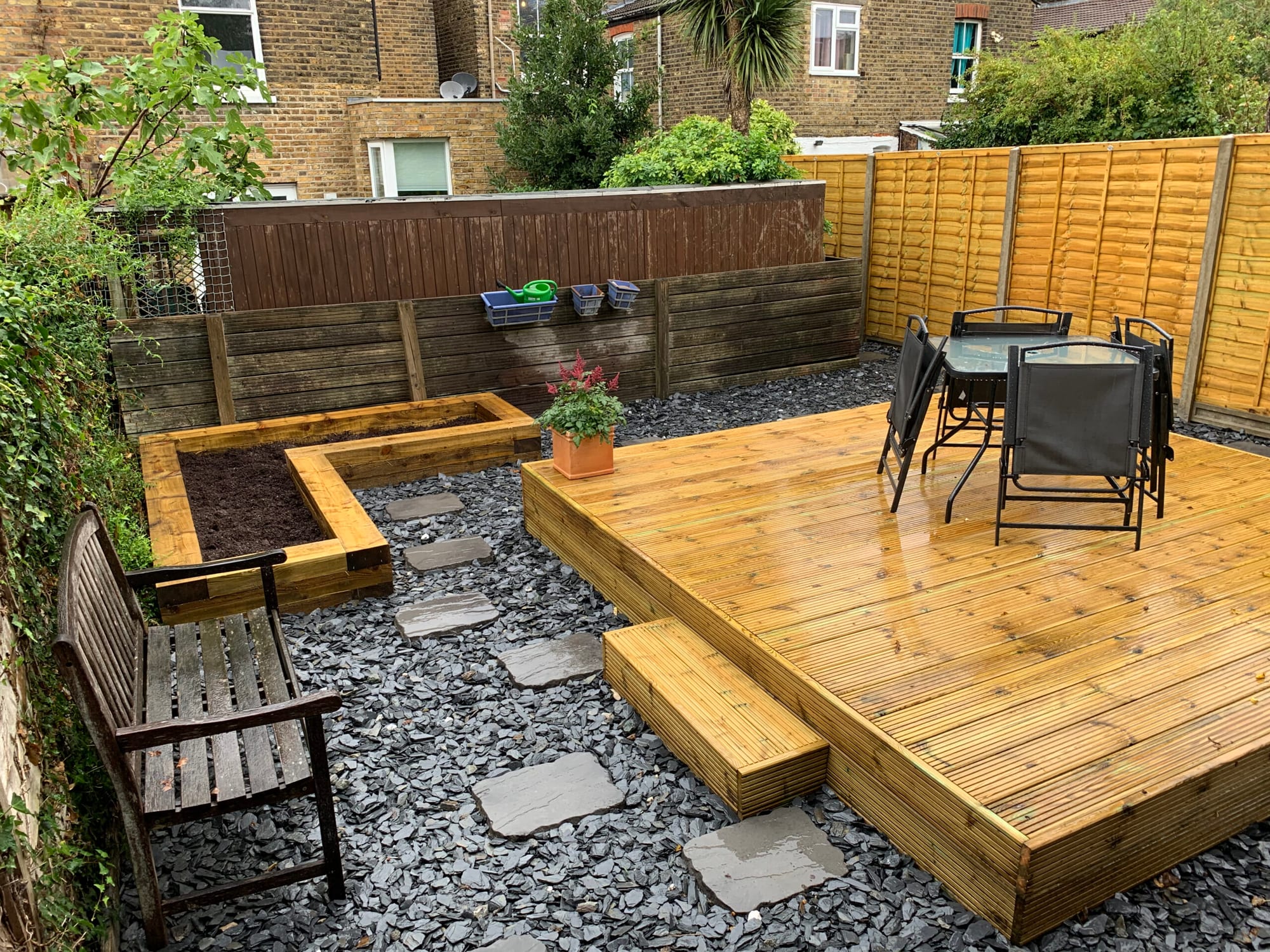 Photo of a landscaped garden with raised timber planter beds filled with soil, a slate chipping path with stepping stones, a wooden bench, and a raised decked seating area with a table and chairs.